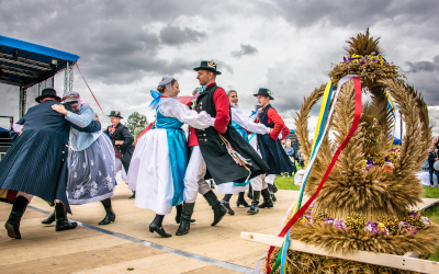 Zdjęcie tańczących ludzi w strojach ludowych. Na pierwszym planie wieniec dożynkowy. Autor zdjęć: Zdjęcie należy do zbiorów Zespołu Folklorystycznego Szamotuły